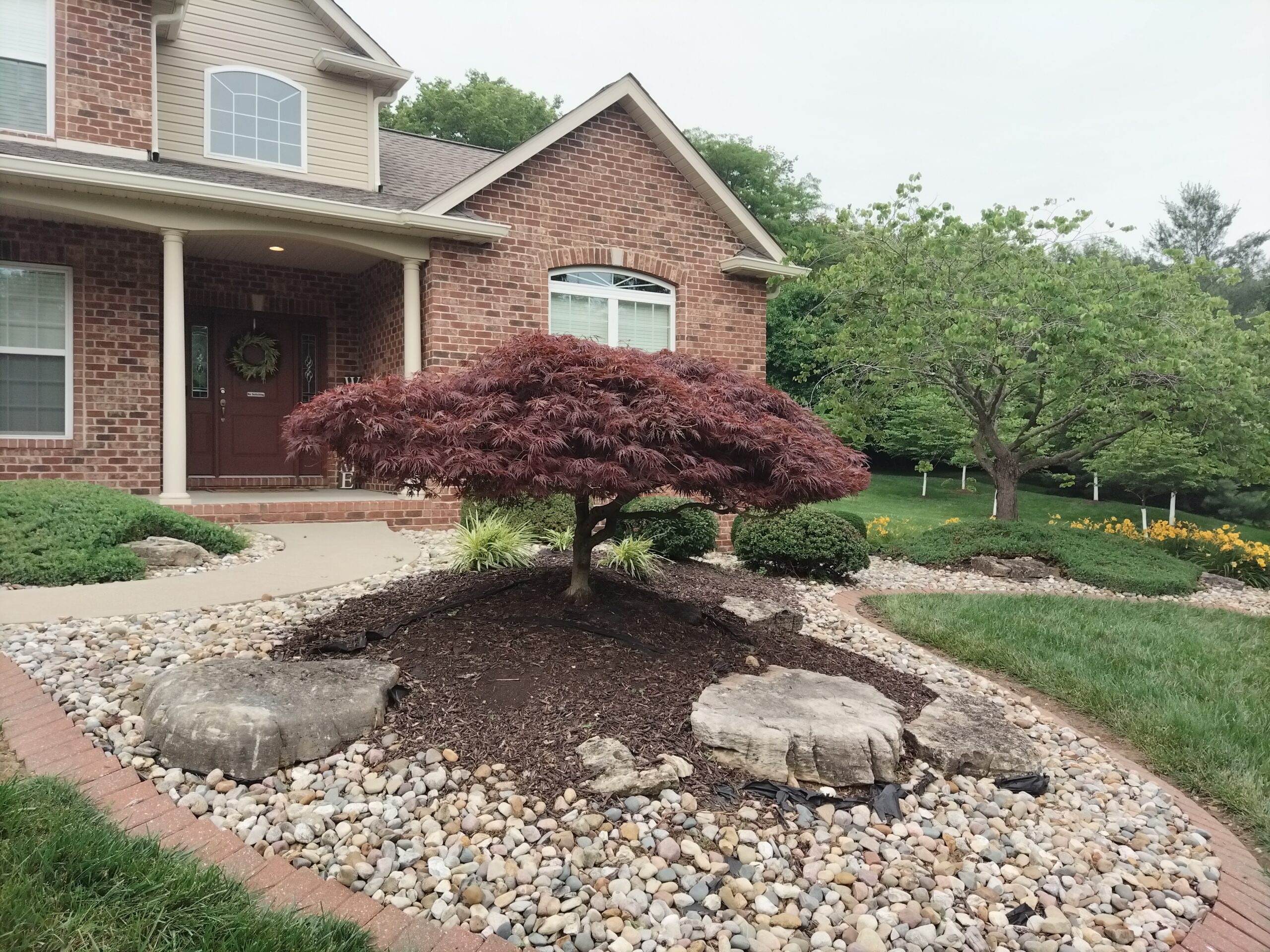 A landscaped area featuring rocks and trees in front of a residential house, showcasing natural elements and greenery.