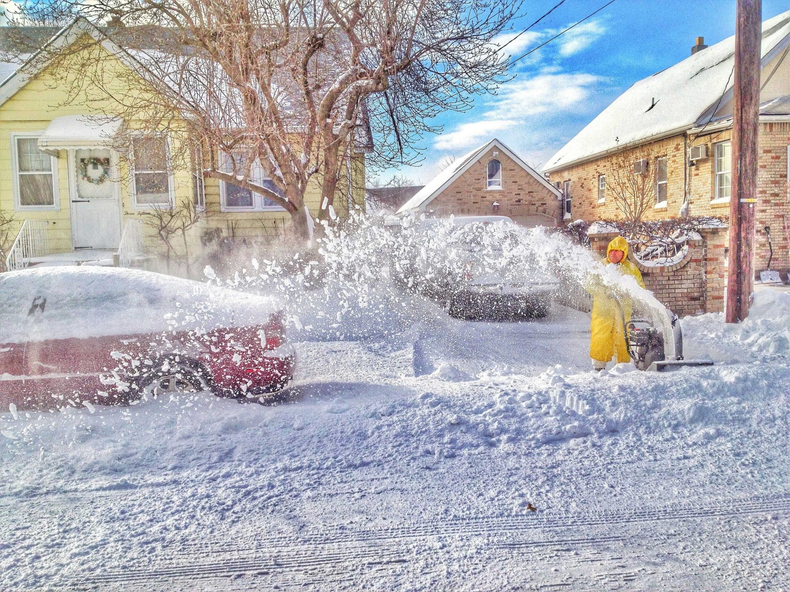 A man in a yellow raincoat blows snow off a car, clearing the windshield in a winter setting.