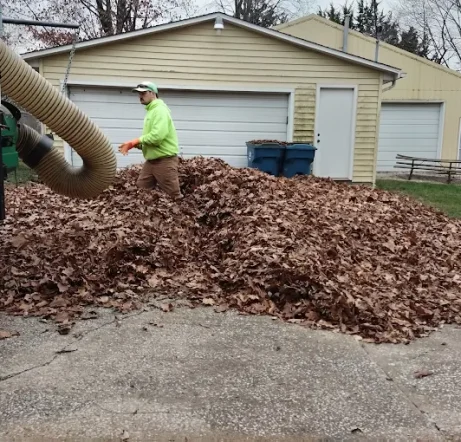 A person in a neon green jacket stands in a large pile of autumn leaves on a driveway, using a vacuum truck. Two garage doors and trees are visible.